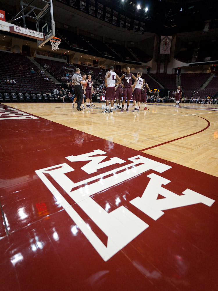 The Texas A&M logo on the edge of the court at Reed Arena with players in background
