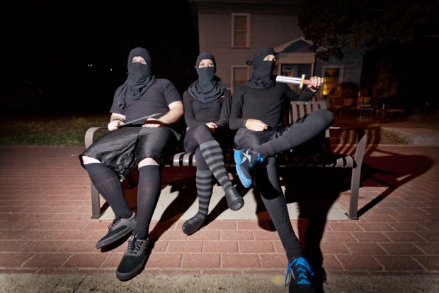 Three college students dressed as ninjas sit on the bench waiting for a bus